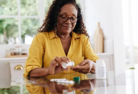 Senior woman in a yellow shirt sitting at a table organizing her medicine - Thrive at Home Network