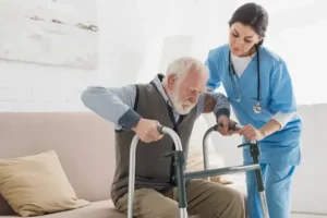Visiting caregiver helping a senior man with a walker up from a sofa. Find these resources and more from Thrive at Home Network